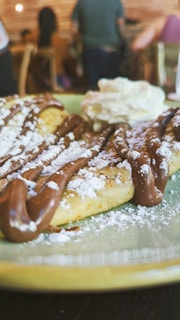 A close-up view of a dessert featuring a plate of waffles drizzled with chocolate sauce and dusted with powdered sugar. There is a dollop of whipped cream on the side. In the background, a blurry interior setting with people seated and wooden furniture.