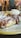 A close-up view of a dessert featuring a plate of waffles drizzled with chocolate sauce and dusted with powdered sugar. There is a dollop of whipped cream on the side. In the background, a blurry interior setting with people seated and wooden furniture.
