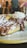 A close-up view of a dessert featuring a plate of waffles drizzled with chocolate sauce and dusted with powdered sugar. There is a dollop of whipped cream on the side. In the background, a blurry interior setting with people seated and wooden furniture.