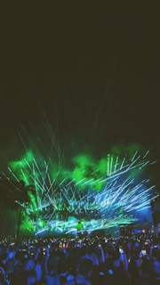 A large crowd gathers under a night sky illuminated by green and blue laser lights. The light beams create dynamic patterns against a backdrop of faint smoke, with silhouettes of palm trees visible. Many people in the crowd are holding up their phones, capturing the event.