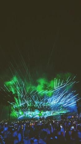 A large crowd gathers under a night sky illuminated by green and blue laser lights. The light beams create dynamic patterns against a backdrop of faint smoke, with silhouettes of palm trees visible. Many people in the crowd are holding up their phones, capturing the event.