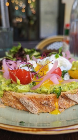 A slice of toasted bread covered with vibrant green avocado spread, topped with bright red cherry tomatoes, thin slices of pickled red onion, and a poached egg with runny yolk. A small salad of mixed greens is placed next to it, and the background includes blurred lights creating a warm ambiance.