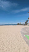 A vast sandy beach with a clear sky above, featuring a row of palm trees in the distance along with distant mountains. A paved path runs along the edge of the sand, with soft shadows cast by the sun.