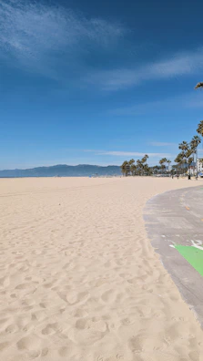A vast sandy beach with a clear sky above, featuring a row of palm trees in the distance along with distant mountains. A paved path runs along the edge of the sand, with soft shadows cast by the sun.