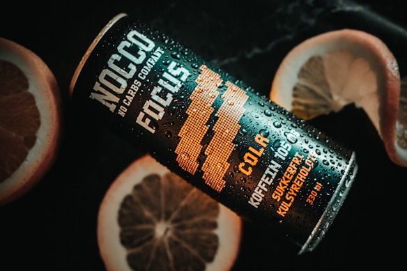 A black and orange can of NOCCO Focus is placed on a dark surface with droplets of water on it, suggesting it is chilled. The can features text such as 'NOCCO NO CARBS COMPANY' and 'COLA' indicating it's a cola-flavored beverage. Slices of grapefruit or similar citrus fruit are positioned around the can, providing a refreshing and vibrant contrast.