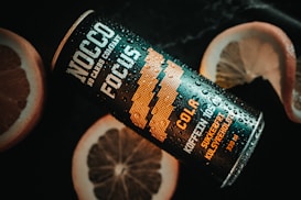 A black and orange can of NOCCO Focus is placed on a dark surface with droplets of water on it, suggesting it is chilled. The can features text such as 'NOCCO NO CARBS COMPANY' and 'COLA' indicating it's a cola-flavored beverage. Slices of grapefruit or similar citrus fruit are positioned around the can, providing a refreshing and vibrant contrast.