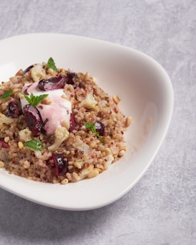 A dish featuring a salad composed of quinoa mixed with chopped nuts, cauli florets, and black olive slices. Topped with a dollop of pink cream or yogurt and garnished with fresh mint leaves, presented in a white, modern, curved bowl, all set against a gray textured background.