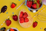 A vibrant display of fresh strawberries arranged on a decorative yellow background. Some strawberries are placed in a heart-shaped arrangement on top of creamy swirls. A metallic cup filled with strawberries is also present, surrounded by scattered colorful gems, a whisk, and a spoon dipped in chocolate. A striped straw adds a playful touch to the scene.