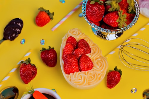 A vibrant display of fresh strawberries arranged on a decorative yellow background. Some strawberries are placed in a heart-shaped arrangement on top of creamy swirls. A metallic cup filled with strawberries is also present, surrounded by scattered colorful gems, a whisk, and a spoon dipped in chocolate. A striped straw adds a playful touch to the scene.