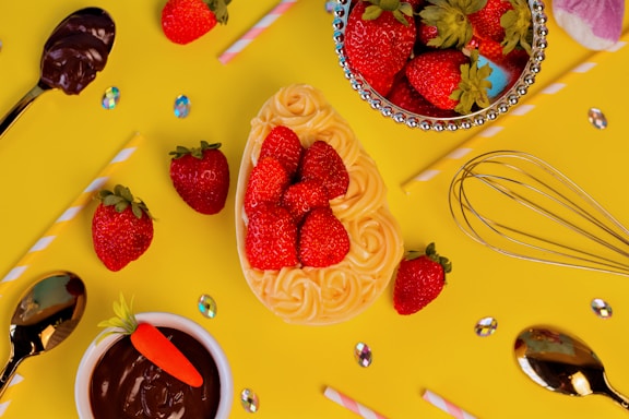 A vibrant arrangement of strawberries placed on top of a cream swirl cake, surrounded by a variety of kitchen utensils including a whisk and spoons with chocolate. The background is a bright yellow with scattered iridescent gems and colorful straws.