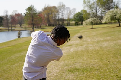 A person with dark hair wearing a white shirt is swinging a golf club on a grassy area near a small body of water. The background features trees with different colored leaves, indicating a park or golf course setting.