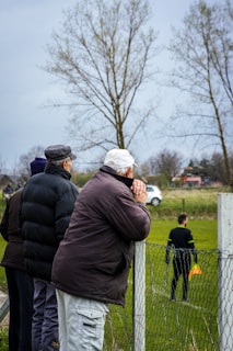 Several older men in heavy jackets and hats are leaning on a wire fence, observing a soccer game in a grassy field. In the background, a referee is standing on the field, holding a yellow flag. Leafless trees and a cloudy sky suggest it is a chilly day.