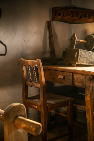 A rustic, dimly lit room featuring a wooden chair and desk. The furniture has a vintage appearance with a slightly distressed finish. A spindle and other woodworking tools are visible, suggesting the room might be a workshop or studio. The walls are bare with subtle cracks, adding to the aged atmosphere.