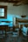 A rustic interior with wooden furniture including a table, chairs, a bed, and a suspended cradle. The walls are made of rough, whitewashed brick, and a window with a wooden frame and iron bars allows in soft light. The atmosphere is serene, with a vintage feel.