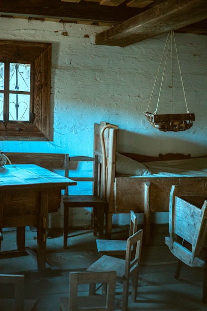 A rustic interior with wooden furniture including a table, chairs, a bed, and a suspended cradle. The walls are made of rough, whitewashed brick, and a window with a wooden frame and iron bars allows in soft light. The atmosphere is serene, with a vintage feel.