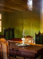 A vintage interior featuring a wooden table with an old-fashioned oil lamp placed on a round tray. The room has wooden chairs with woven backs, and green walls with a warm, rustic feel. The lighting casts soft shadows, contributing to a cozy and nostalgic atmosphere.