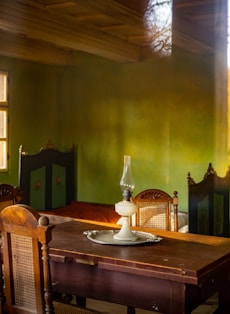 A vintage interior featuring a wooden table with an old-fashioned oil lamp placed on a round tray. The room has wooden chairs with woven backs, and green walls with a warm, rustic feel. The lighting casts soft shadows, contributing to a cozy and nostalgic atmosphere.
