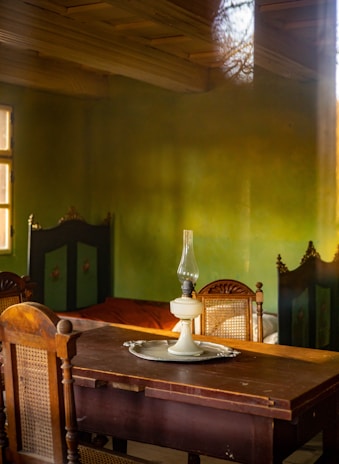 A vintage interior featuring a wooden table with an old-fashioned oil lamp placed on a round tray. The room has wooden chairs with woven backs, and green walls with a warm, rustic feel. The lighting casts soft shadows, contributing to a cozy and nostalgic atmosphere.