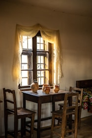 A rustic interior scene featuring a wooden table with four wooden chairs. On the table, there are several clay pots and jugs. A window behind the table is adorned with sheer curtains, allowing soft light to filter in, creating a warm, inviting atmosphere. A colorful woven blanket adds detail to a bench in the background.