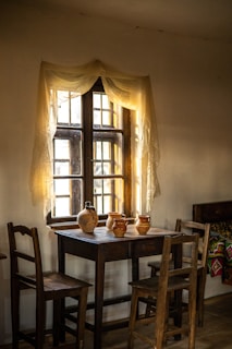 A rustic interior scene featuring a wooden table with four wooden chairs. On the table, there are several clay pots and jugs. A window behind the table is adorned with sheer curtains, allowing soft light to filter in, creating a warm, inviting atmosphere. A colorful woven blanket adds detail to a bench in the background.