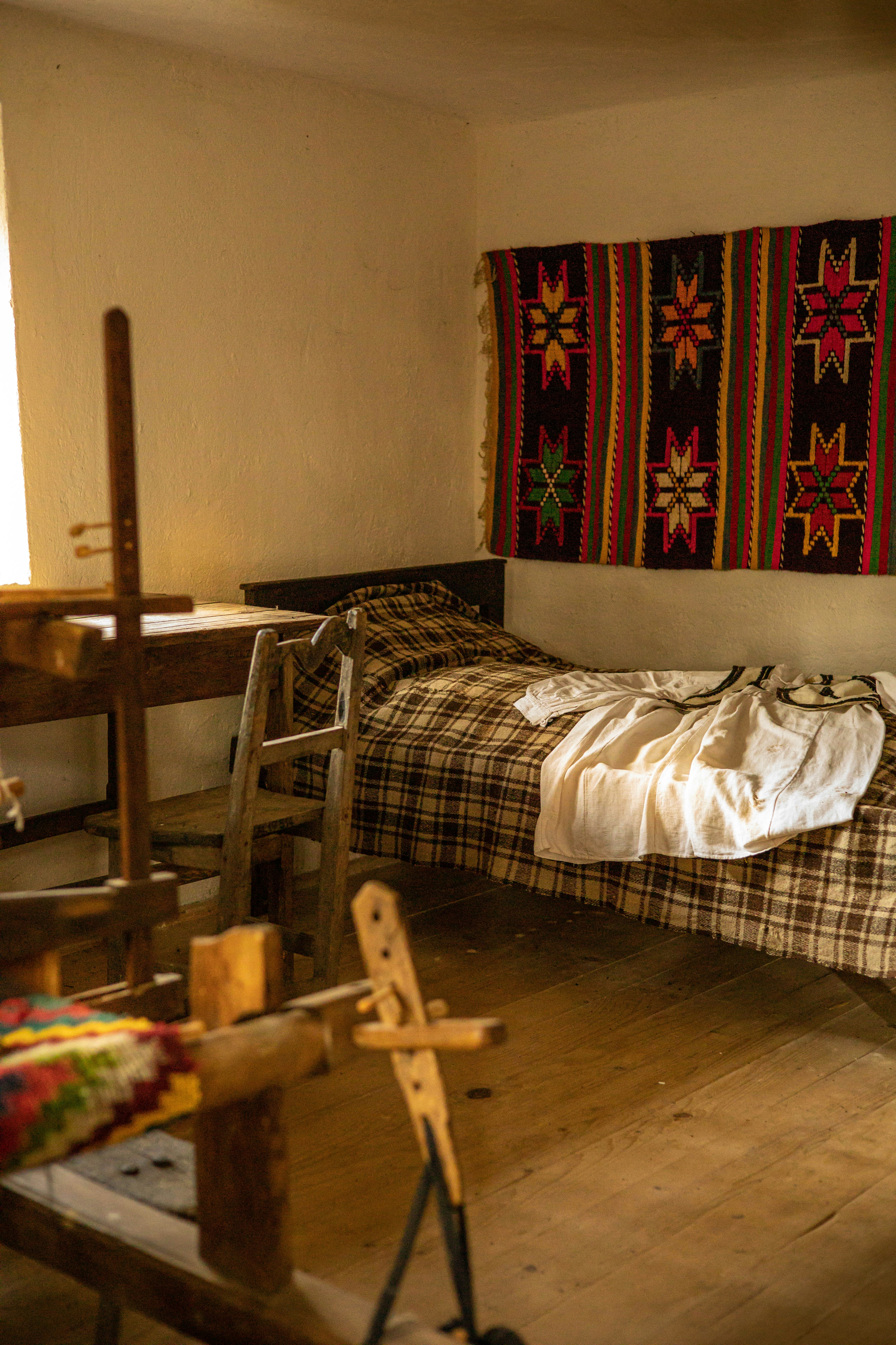 A cozy room features a wooden loom in the foreground, a simple bed with a plaid blanket, and a vibrant handwoven textile with star patterns hanging on the wall. Soft, natural light filters in from a window, illuminating the rustic wooden furniture and floor.