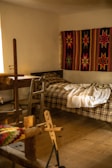 A cozy room features a wooden loom in the foreground, a simple bed with a plaid blanket, and a vibrant handwoven textile with star patterns hanging on the wall. Soft, natural light filters in from a window, illuminating the rustic wooden furniture and floor.