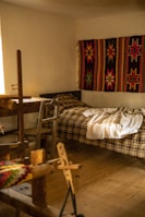 A cozy room features a wooden loom in the foreground, a simple bed with a plaid blanket, and a vibrant handwoven textile with star patterns hanging on the wall. Soft, natural light filters in from a window, illuminating the rustic wooden furniture and floor.
