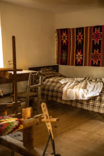 A cozy room features a wooden loom in the foreground, a simple bed with a plaid blanket, and a vibrant handwoven textile with star patterns hanging on the wall. Soft, natural light filters in from a window, illuminating the rustic wooden furniture and floor.