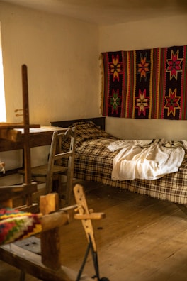 A cozy room features a wooden loom in the foreground, a simple bed with a plaid blanket, and a vibrant handwoven textile with star patterns hanging on the wall. Soft, natural light filters in from a window, illuminating the rustic wooden furniture and floor.
