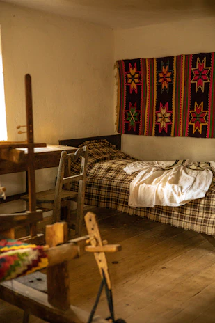 A cozy room features a wooden loom in the foreground, a simple bed with a plaid blanket, and a vibrant handwoven textile with star patterns hanging on the wall. Soft, natural light filters in from a window, illuminating the rustic wooden furniture and floor.