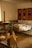 A cozy room features a wooden loom in the foreground, a simple bed with a plaid blanket, and a vibrant handwoven textile with star patterns hanging on the wall. Soft, natural light filters in from a window, illuminating the rustic wooden furniture and floor.