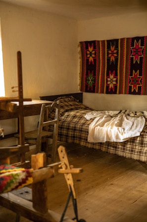 A cozy room features a wooden loom in the foreground, a simple bed with a plaid blanket, and a vibrant handwoven textile with star patterns hanging on the wall. Soft, natural light filters in from a window, illuminating the rustic wooden furniture and floor.