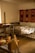 A cozy room features a wooden loom in the foreground, a simple bed with a plaid blanket, and a vibrant handwoven textile with star patterns hanging on the wall. Soft, natural light filters in from a window, illuminating the rustic wooden furniture and floor.