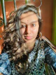 A person with long, wavy hair wearing a blue patterned garment sits in sunlight against a decorative metal structure. A red bindi is present on the forehead.