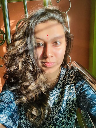 A person with long, wavy hair wearing a blue patterned garment sits in sunlight against a decorative metal structure. A red bindi is present on the forehead.