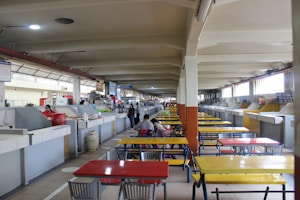 A spacious indoor food market with rows of stalls and colorful tables. The tables are painted in bright red, yellow, and blue. A few people are present, and some stalls appear open with items displayed.