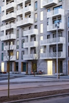 A modern building featuring multiple balconies with white railings and a facade of light gray bricks. The windows are tall and narrow, and several bicycles are parked near the entrance. Sparse trees are planted along a paved sidewalk.
