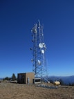 A tall telecommunications tower is equipped with various antennas and satellite dishes, located on a rocky terrain with sparse vegetation. A small building is situated beside the tower, likely housing equipment. The background features a clear blue sky and distant mountain ranges.