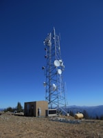 A tall telecommunications tower is equipped with various antennas and satellite dishes, located on a rocky terrain with sparse vegetation. A small building is situated beside the tower, likely housing equipment. The background features a clear blue sky and distant mountain ranges.