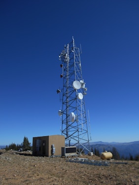 A tall telecommunications tower is equipped with various antennas and satellite dishes, located on a rocky terrain with sparse vegetation. A small building is situated beside the tower, likely housing equipment. The background features a clear blue sky and distant mountain ranges.