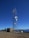 A tall telecommunications tower is equipped with various antennas and satellite dishes, located on a rocky terrain with sparse vegetation. A small building is situated beside the tower, likely housing equipment. The background features a clear blue sky and distant mountain ranges.