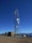 A tall telecommunications tower is equipped with various antennas and satellite dishes, located on a rocky terrain with sparse vegetation. A small building is situated beside the tower, likely housing equipment. The background features a clear blue sky and distant mountain ranges.