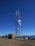 A tall telecommunications tower is equipped with various antennas and satellite dishes, located on a rocky terrain with sparse vegetation. A small building is situated beside the tower, likely housing equipment. The background features a clear blue sky and distant mountain ranges.
