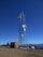 A tall telecommunications tower is equipped with various antennas and satellite dishes, located on a rocky terrain with sparse vegetation. A small building is situated beside the tower, likely housing equipment. The background features a clear blue sky and distant mountain ranges.