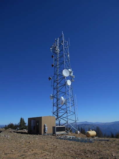 A tall telecommunications tower is equipped with various antennas and satellite dishes, located on a rocky terrain with sparse vegetation. A small building is situated beside the tower, likely housing equipment. The background features a clear blue sky and distant mountain ranges.