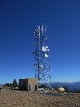 A tall telecommunications tower is equipped with various antennas and satellite dishes, located on a rocky terrain with sparse vegetation. A small building is situated beside the tower, likely housing equipment. The background features a clear blue sky and distant mountain ranges.