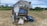 An agricultural machine is situated in a harvested rice field, surrounded by dry straw and a stack of blue tarpaulins. The machine appears to be a thresher, with various mechanisms exposed. In the background, banana trees and other green vegetation are visible, along with a clear blue sky.