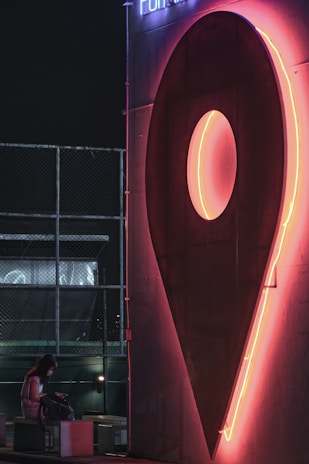 A large neon sign in the shape of a navigation pin glows brightly in pink and orange hues against a dark backdrop. Below the sign, a person is seated on a bench, looking into a bag. The setting appears to be urban, possibly a rooftop, with a chain-link fence visible in the background.