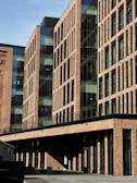 A modern brick building with large glass windows and a linear architectural design. The facade displays a repetitive pattern of rectangular windows, creating a blend of traditional and contemporary styles. The scene is well-lit with shadows suggesting a sunny day.