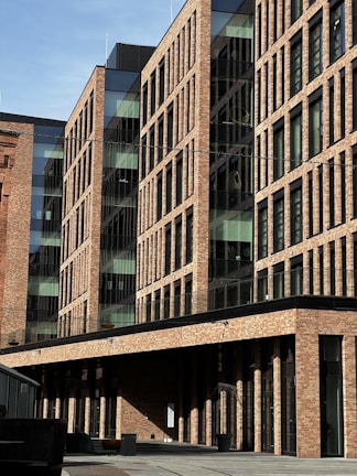 A modern brick building with large glass windows and a linear architectural design. The facade displays a repetitive pattern of rectangular windows, creating a blend of traditional and contemporary styles. The scene is well-lit with shadows suggesting a sunny day.